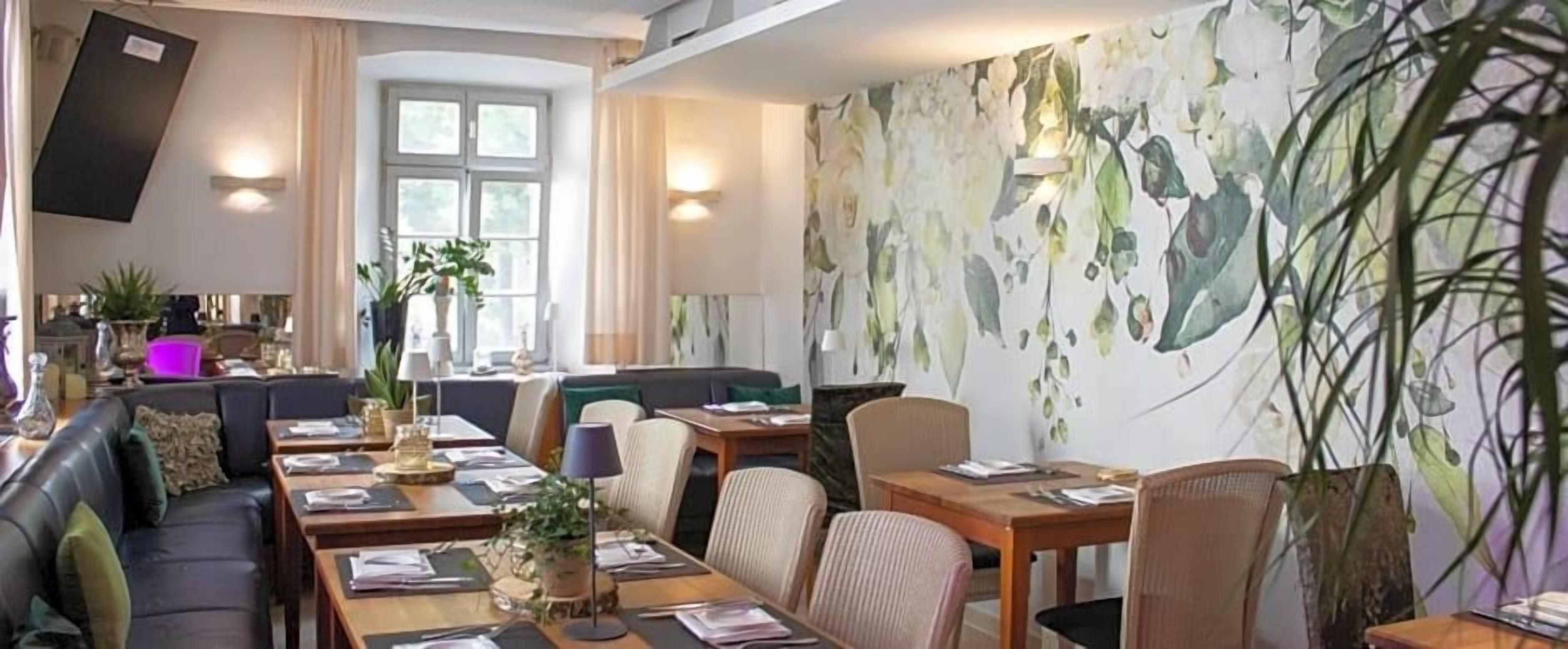 A dining area with a long table, chairs, and a sofa. There are plants and a wall with a floral mural.