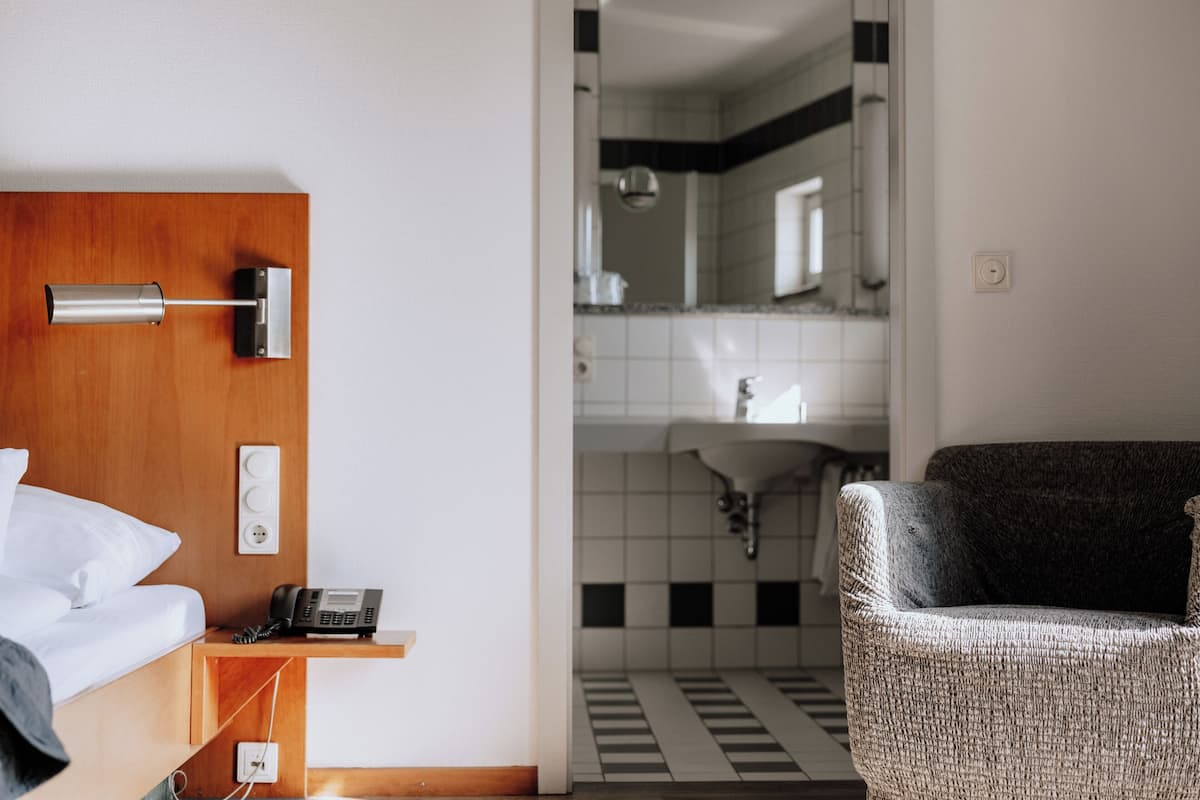 Ein Hotelzimmer mit einem hölzernen Kopfteil, einem Nachttischtelefon, einem kleinen Tisch, einem Sessel und Blick ins Badezimmer mit gefliestem Boden und Waschbecken.
