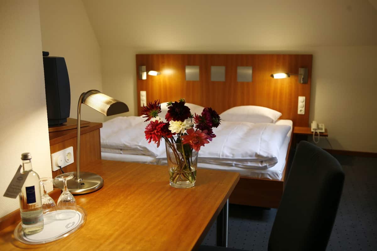 Ein Hotelzimmer mit Schreibtisch aus Holz, einem Nachttisch, einer Lampe, einer Vase mit Blumen, einer Flasche und einem Glas.