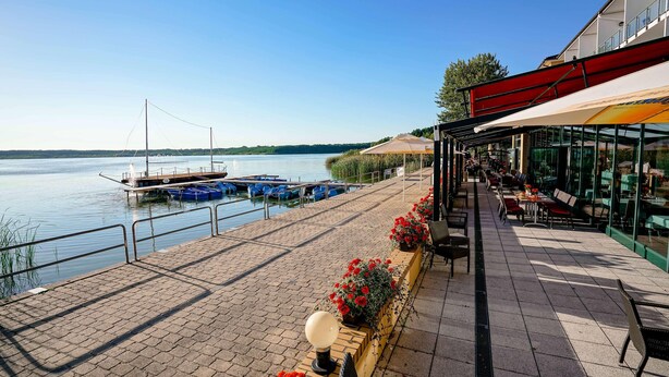 A waterfront patio with outdoor seating, a dock with boats, and a restaurant with a glass facade. - TOP Countryline Seebad Casino (Rangsdorf)