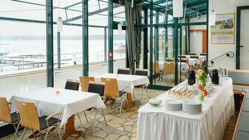 A restaurant with tables set for dining, large windows offering a view of the water, and a buffet table with food and drinks.