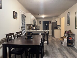 A dining area with a dark wooden table and chairs, a kitchen in the background with white cabinets, and a cat on a shelf. - Special Introductory Price: 5 Bedroom Home. Safe, Great location & Amenities, (Fairburn)