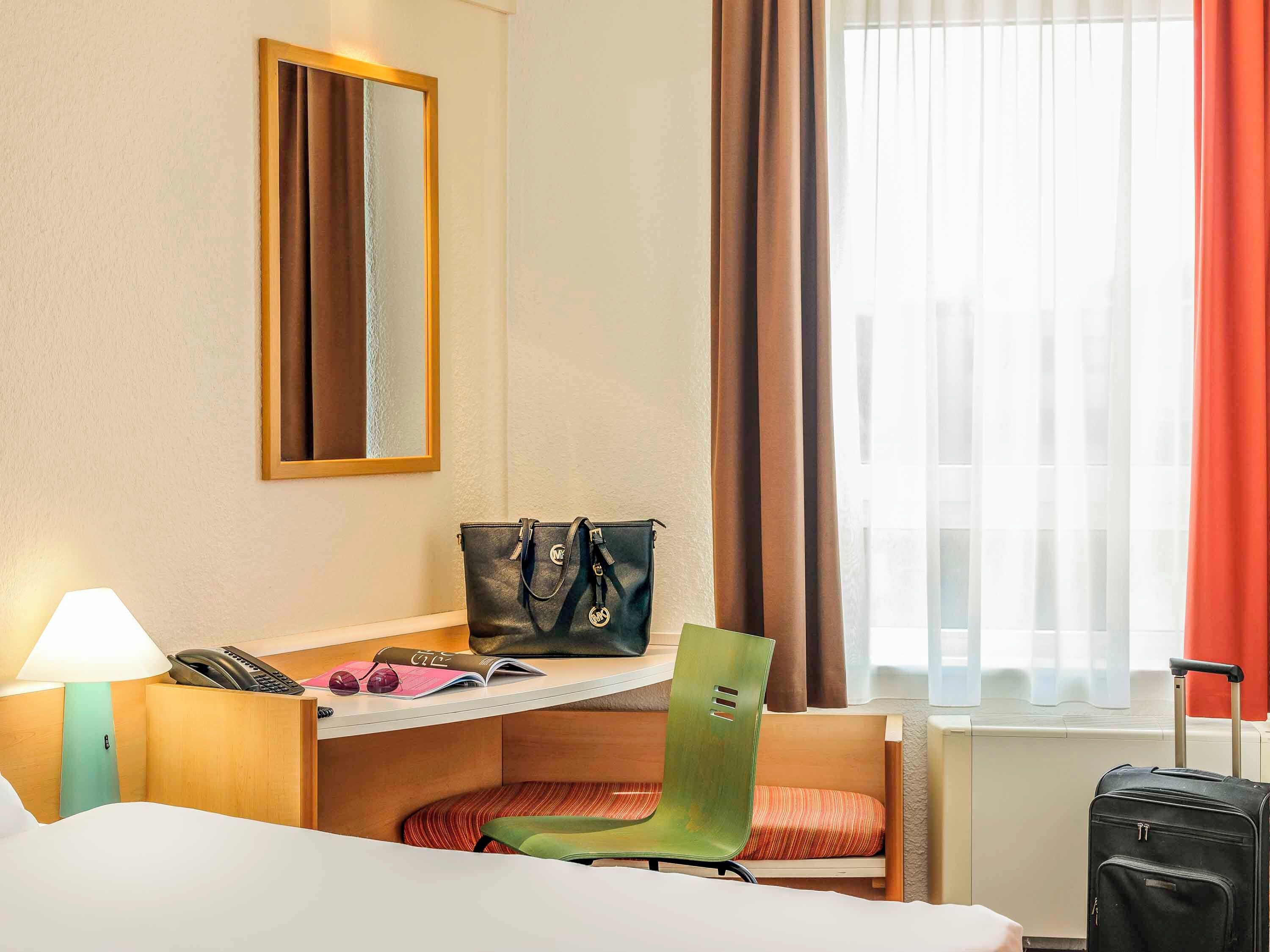 A hotel room with a bed, a desk with a lamp, a chair, a suitcase, and a window with curtains.