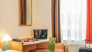 A hotel room with a bed, a desk with a lamp, a chair, a suitcase, and a window with curtains.