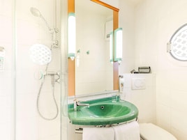 Baño con ducha envuelta en vidrio, lavabo verde, inodoro blanco y espejo con marco de madera.