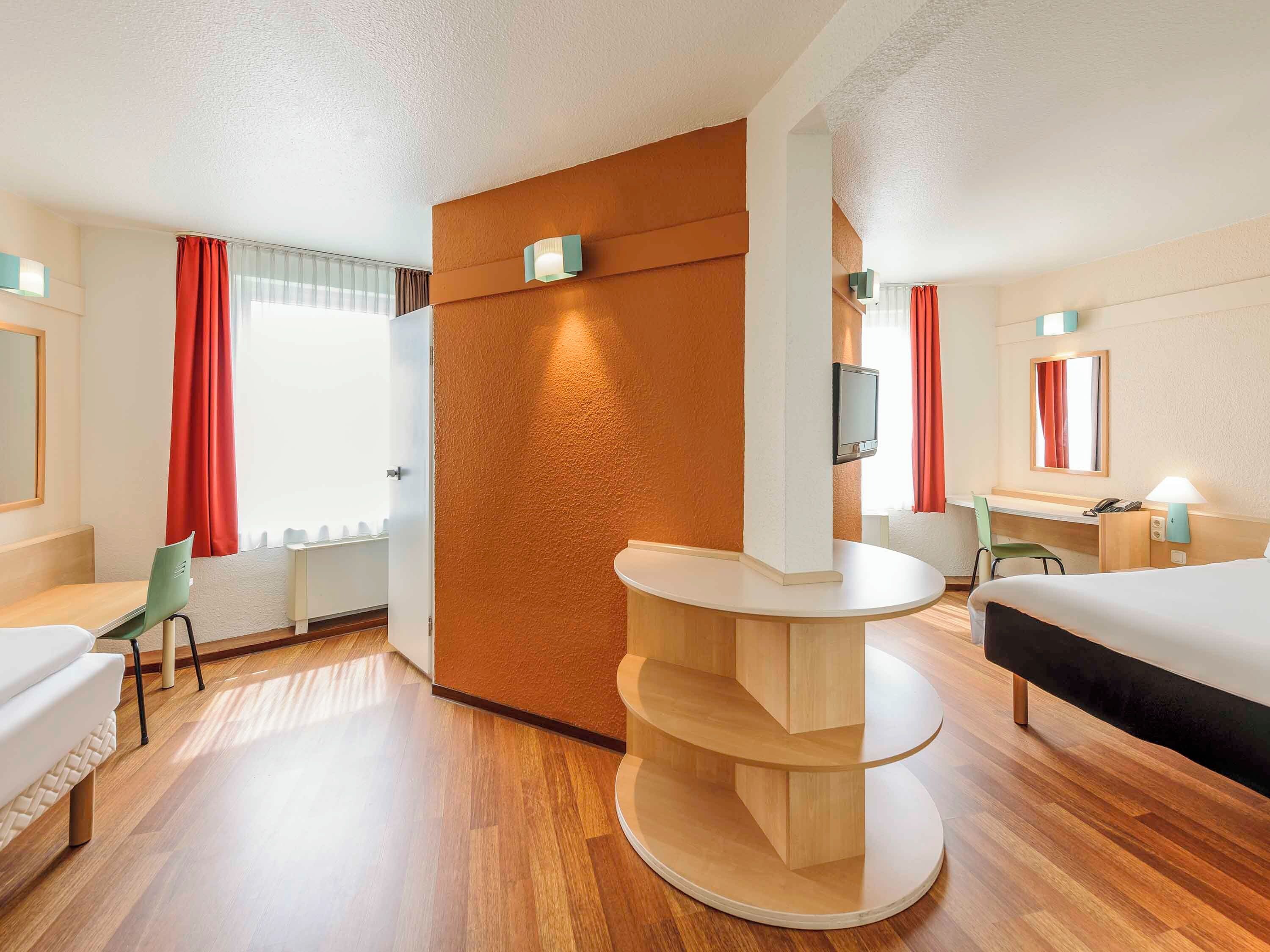 A hotel room with a bed, a desk, a chair, a round wooden table, and a TV mounted on the wall.