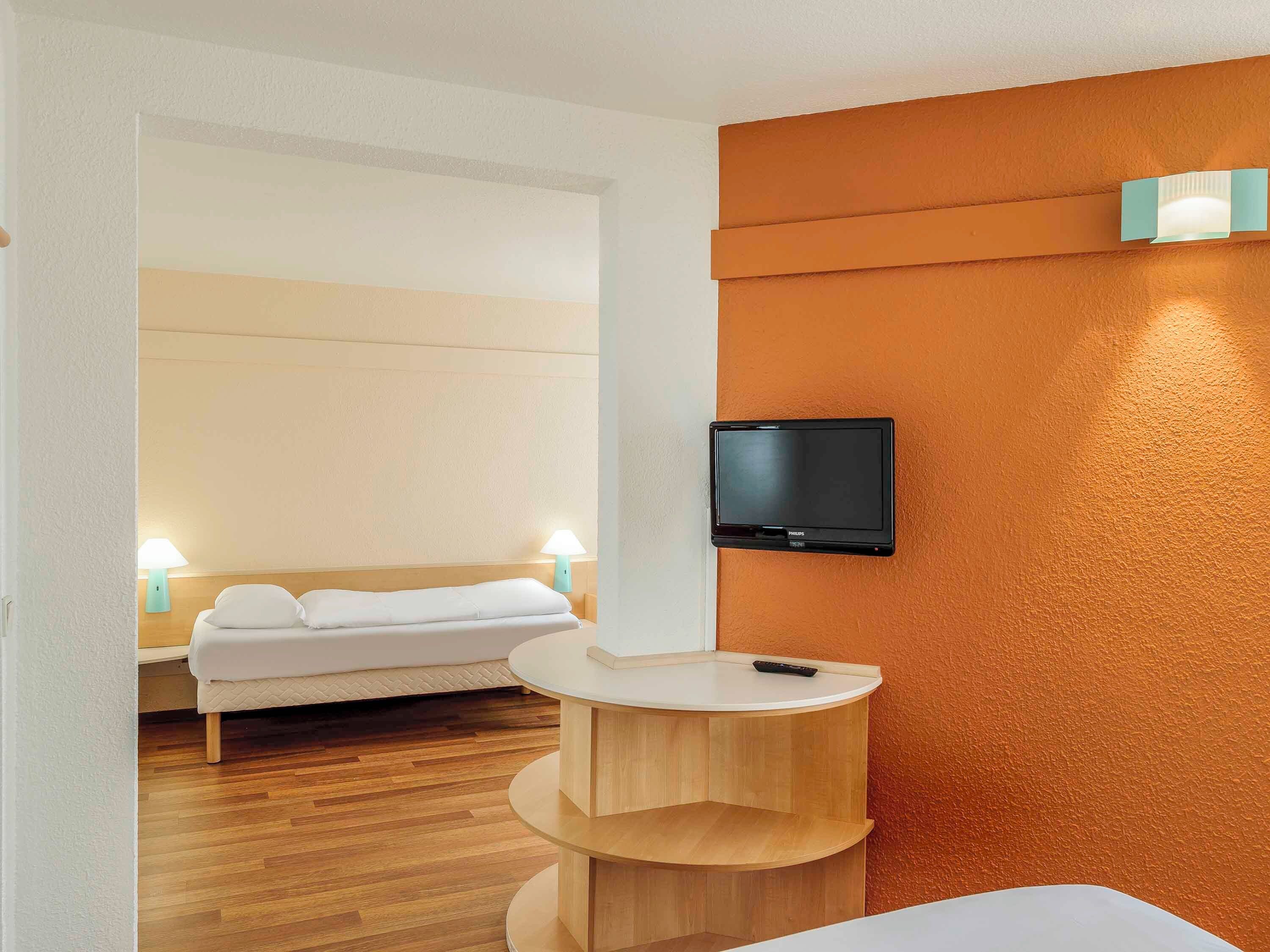 A hotel room with a bed, a TV mounted on the wall, a small wooden table, and a lamp.