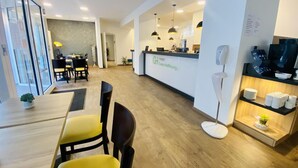 Área de recepção de hotel, com balcão, espaço de espera com cadeiras amarelas, abajur e televisão em uma estante.