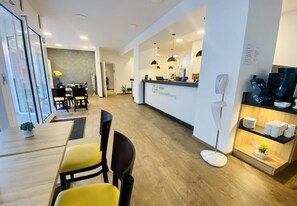 Un espace d’accueil d’hôtel comprenant un comptoir de réception, un coin salon avec des chaises jaunes, un lampadaire et un téléviseur posé sur une étagère.