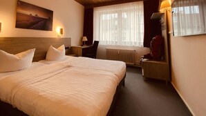 Quarto de hotel com uma cama grande, uma escrivaninha, uma cadeira, um radiador e uma janela com cortinas leves.