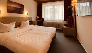 Une chambre d’hôtel avec un grand lit, un bureau, une chaise, un radiateur et une fenêtre avec des rideaux légers.