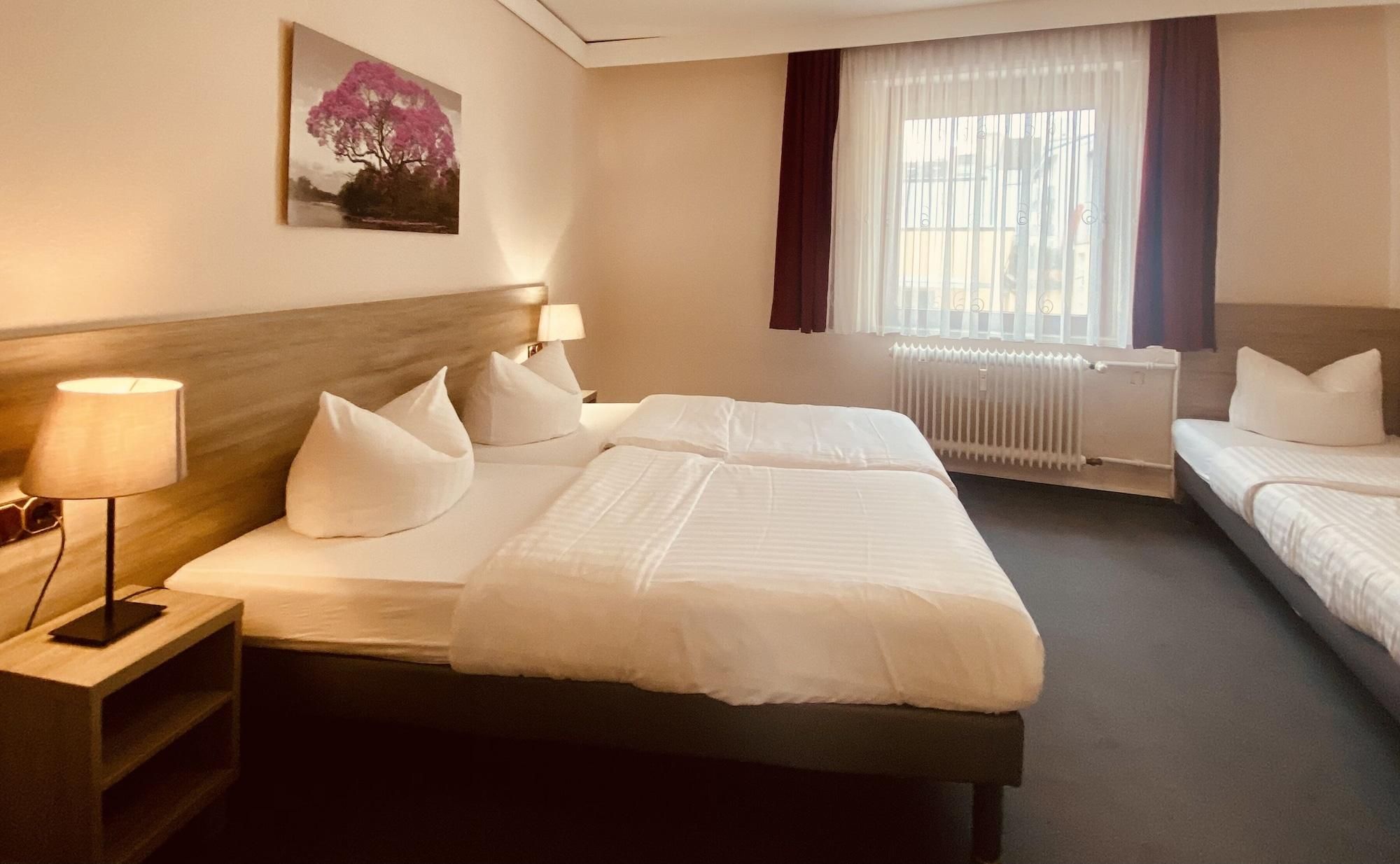 Ein Hotelzimmer mit zwei Betten, einem Nachttisch mit Lampe, einem Bild an der Wand und einem Fenster mit Vorhängen.