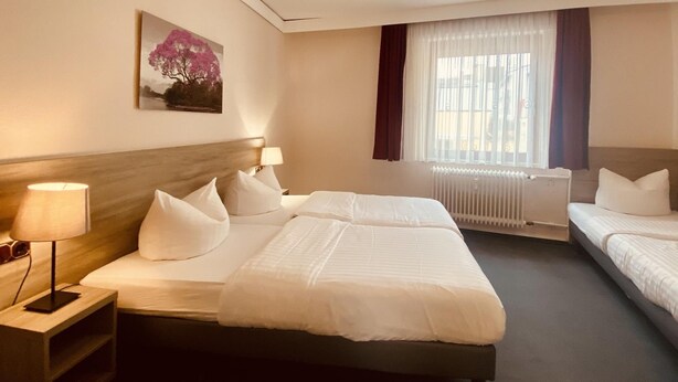 Quarto de hotel com duas camas, mesa de cabeceira com abajur, quadro na parede e janela com cortinas.
