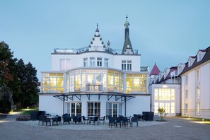 Ein großes, weißes Gebäude mit einer geschwungenen Glasfassade, einem Schild mit der Aufschrift "PARKHOTEL" und einer Außenbereich-Sitzgelegenheit mit Tischen und Stühlen.