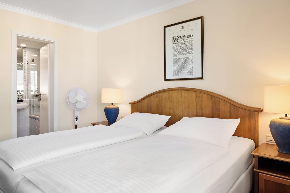 Une chambre d’hôtel avec deux lits, une tête de lit en bois, un ventilateur fixé au mur, une lampe de chevet et un texte encadré accroché au mur.