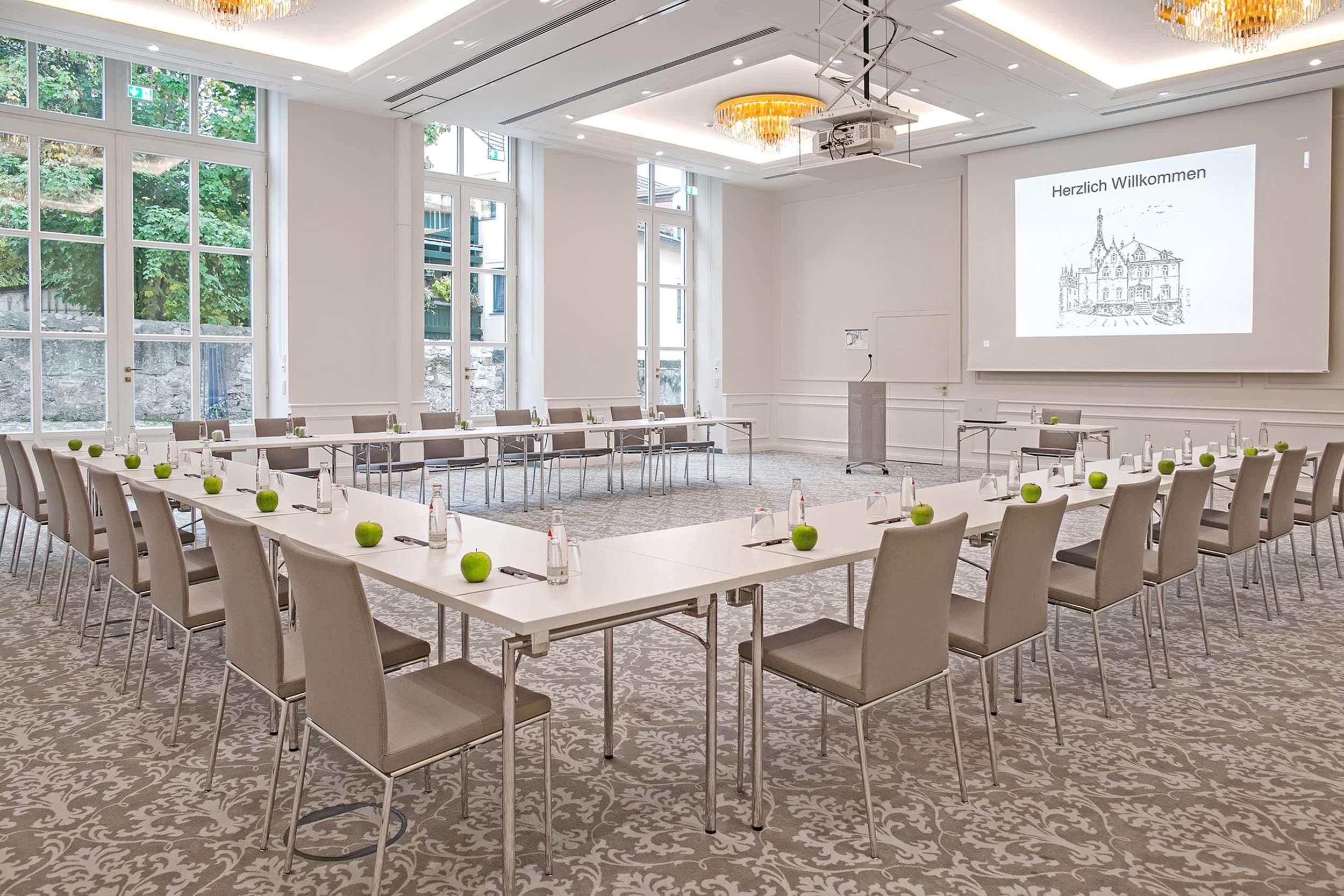 Una sala conferenze con un grande tavolo, sedie, uno schermo per proiettore che mostra un messaggio di benvenuto e ampie finestre con vista su spazi verdi.