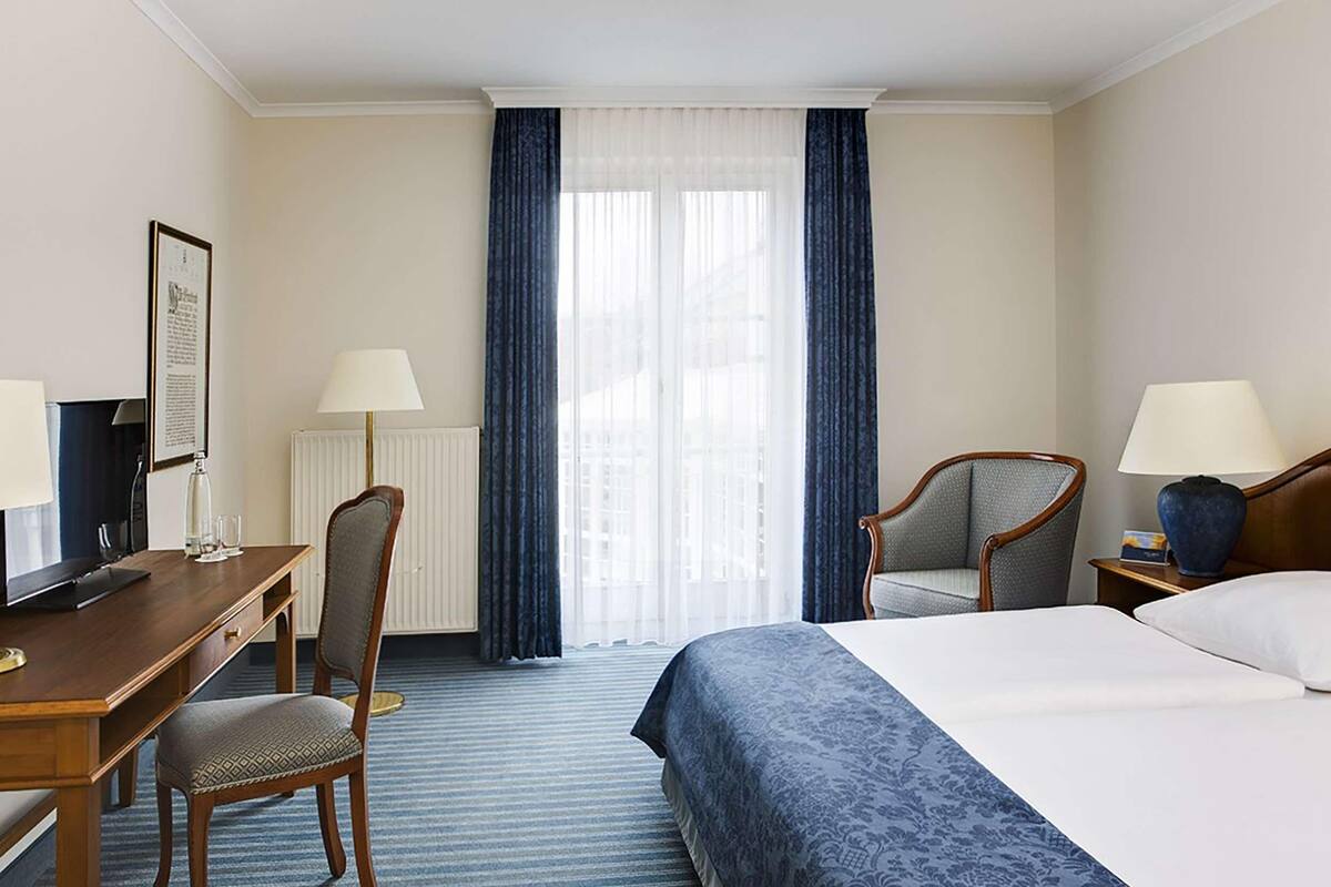 A hotel room with a bed, a desk, a chair, a lamp, and a window with curtains.