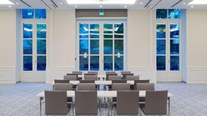 Un salón de conferencias con una gran lámpara de araña, varias mesas y ventanas amplias con vistas a árboles.