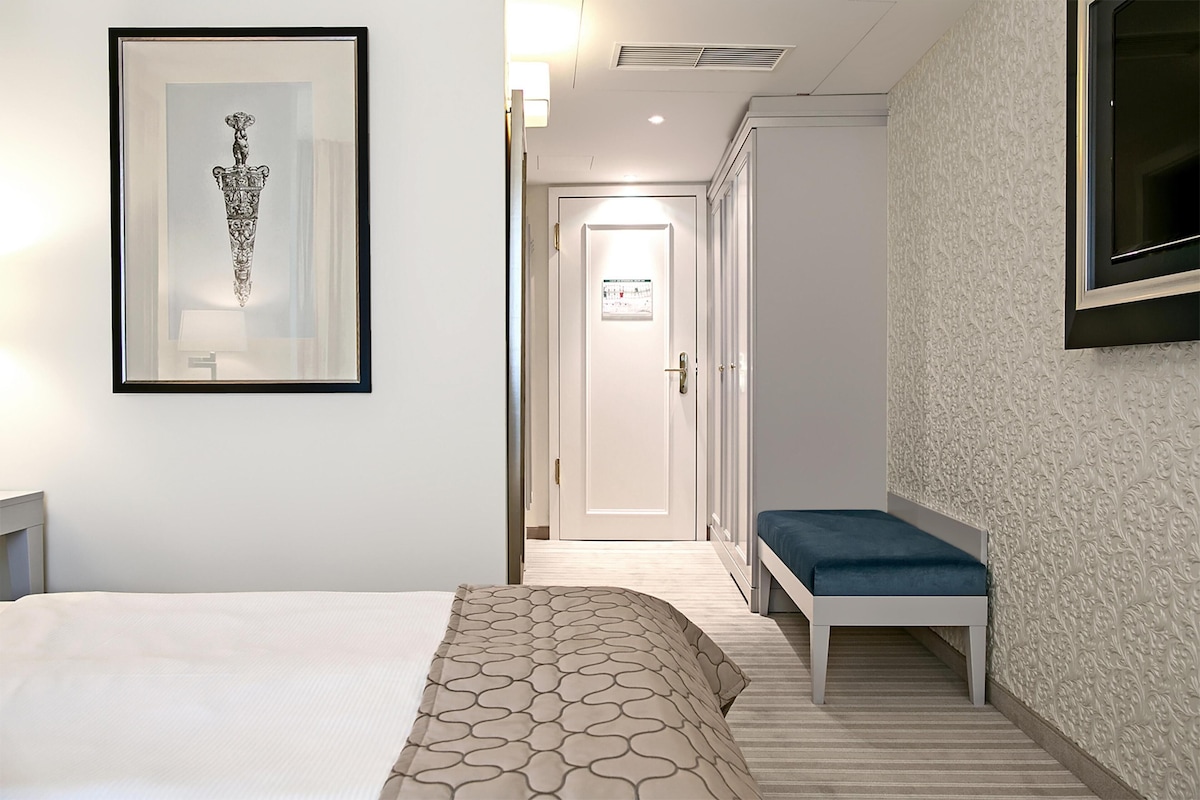 A hotel room with a bed, a framed picture, a bench, and a door leading to another room.