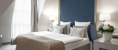 A hotel room with a large bed, two bedside tables, a desk, a chair, a window with curtains, and a wall-mounted lamp.