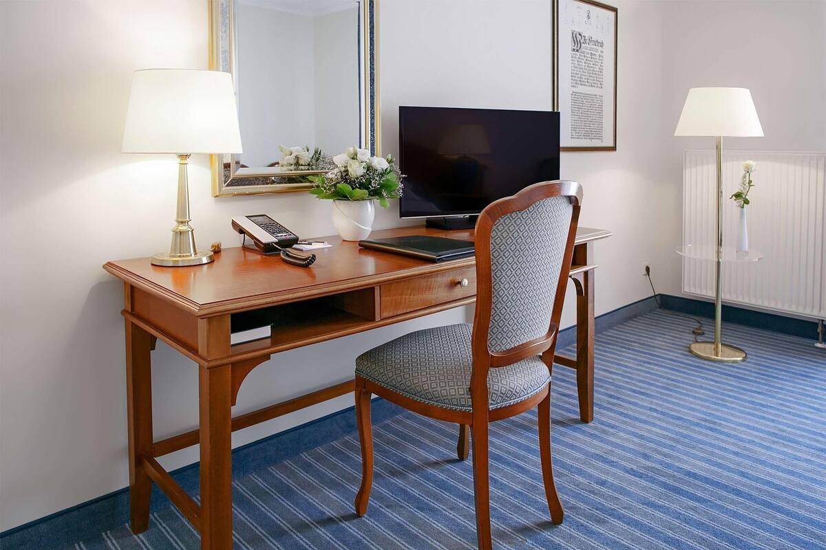 A hotel room with a wooden desk, a chair, a television, a telephone, a vase with flowers, and two lamps.