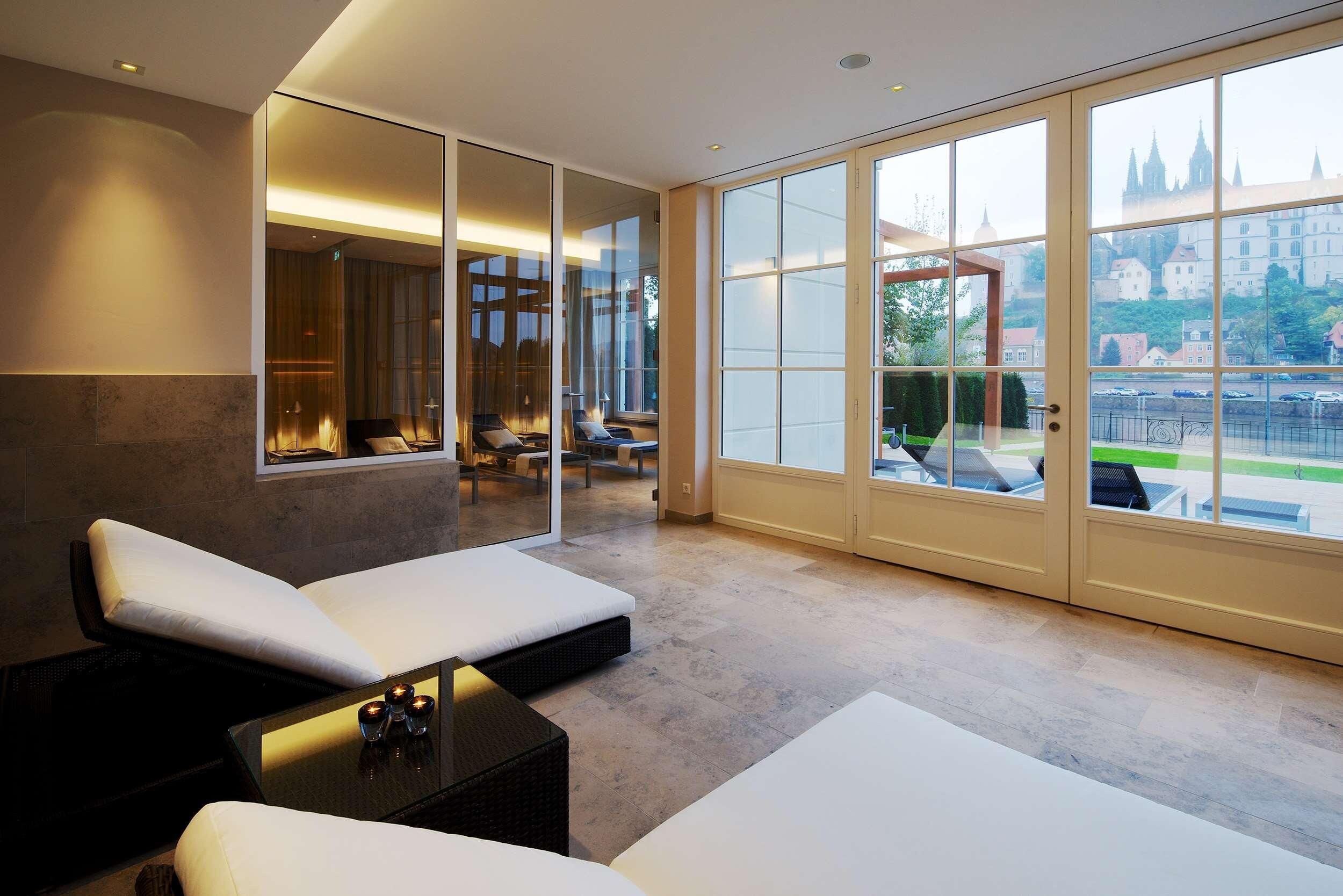 A modern hotel room with a large window, a seating area with white cushions, and a view of a castle.