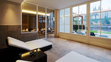 A modern hotel room with a large window, a seating area with white cushions, and a view of a castle.