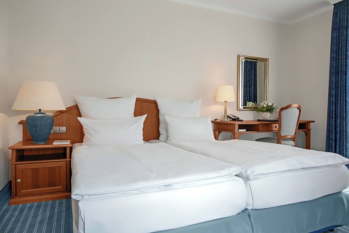 A double bed with white linens, a wooden nightstand with a lamp, a desk with a chair, and a mirror on the wall.