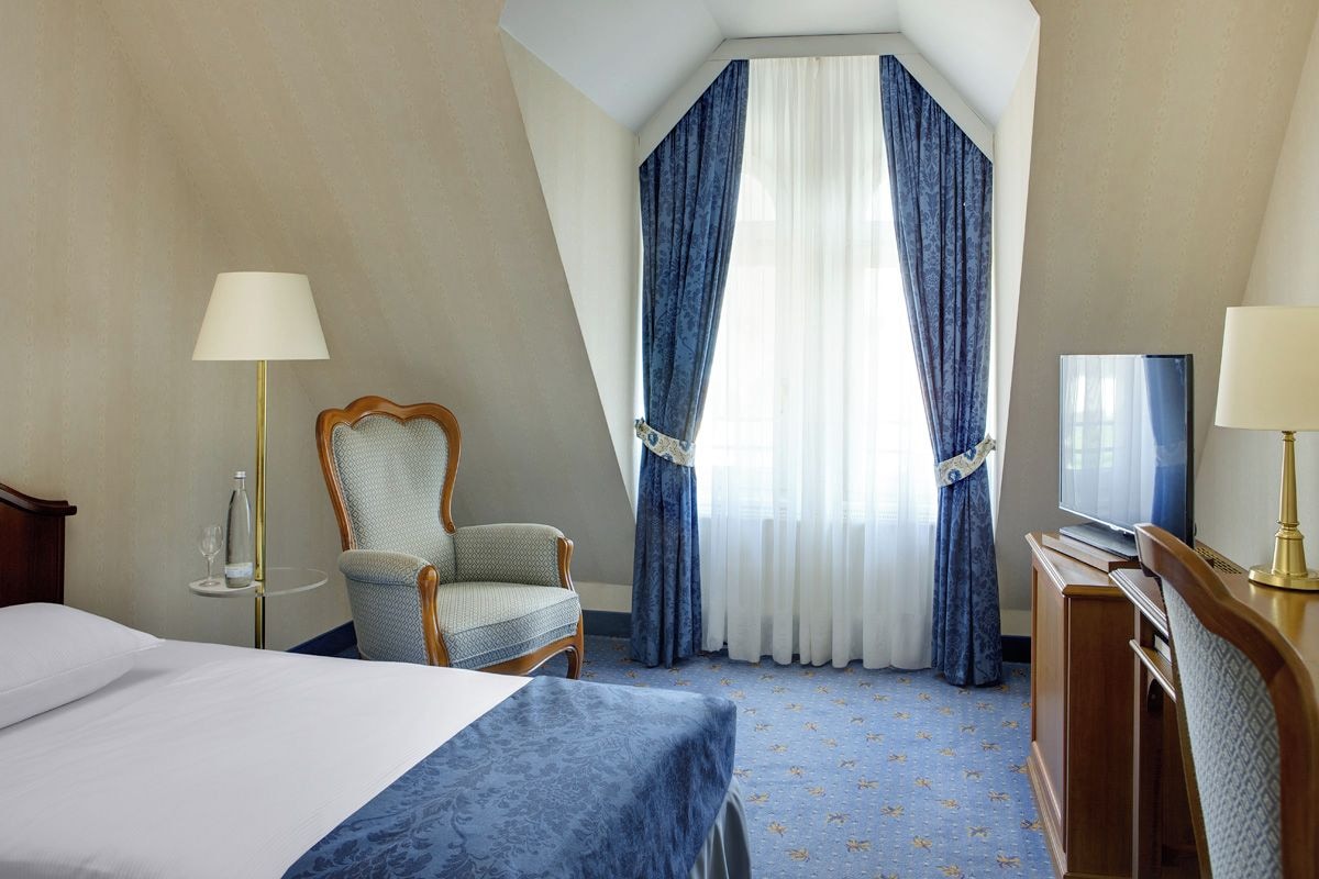 Une chambre d’hôtel avec un lit, une chaise, un bureau avec une télévision et une fenêtre avec des rideaux.