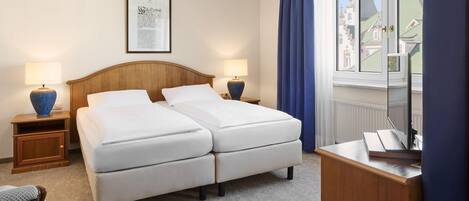 Habitación de hotel con cama doble, dos mesitas de noche con lámparas, un escritorio con silla, un cuadro en la pared y ventana con vistas a edificios.