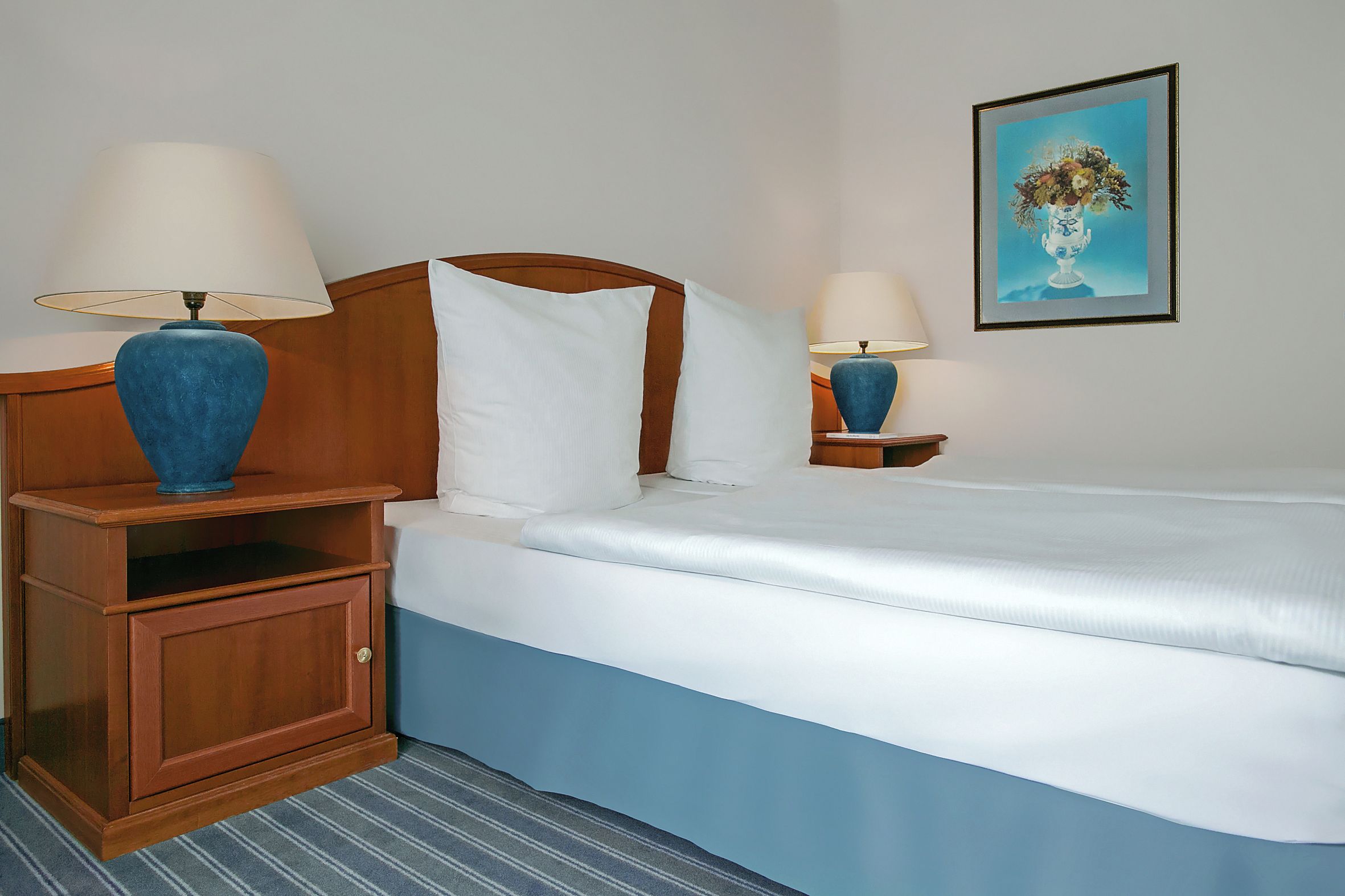 Habitación de hotel con cama, dos mesitas de noche con lámparas, un mueble auxiliar de madera, un cuadro en la pared y una alfombra a rayas.