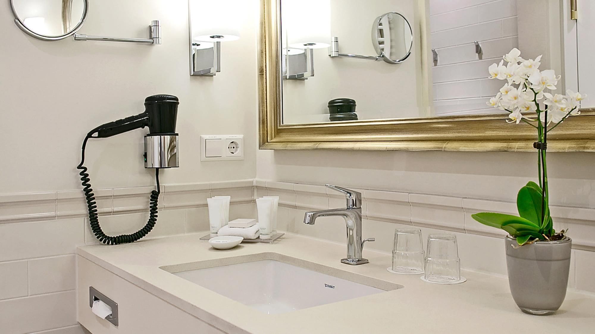 A hotel bathroom with a sink, mirror, hairdryer, and a potted orchid.