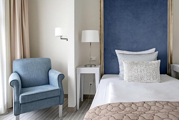 Una habitación de hotel con una cama, un sillón azul, una mesita de noche con una lámpara y una pared con un detalle azul.