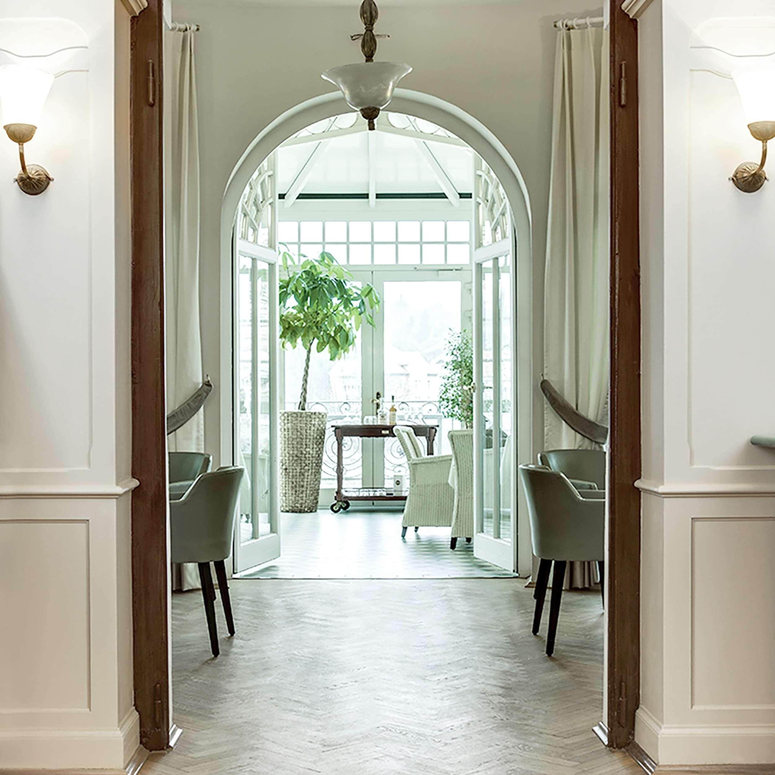 Un pasillo con piso blanco, un amplio arco de entrada y vista a un espacio exterior con mesa y sillas.