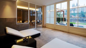 Una habitación de hotel moderna con un ventanal grande, una zona de estar con cojines blancos y vistas a un castillo.