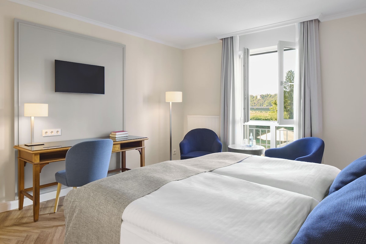 A hotel room with a bed, two chairs, a desk, a TV, and a window with a view.