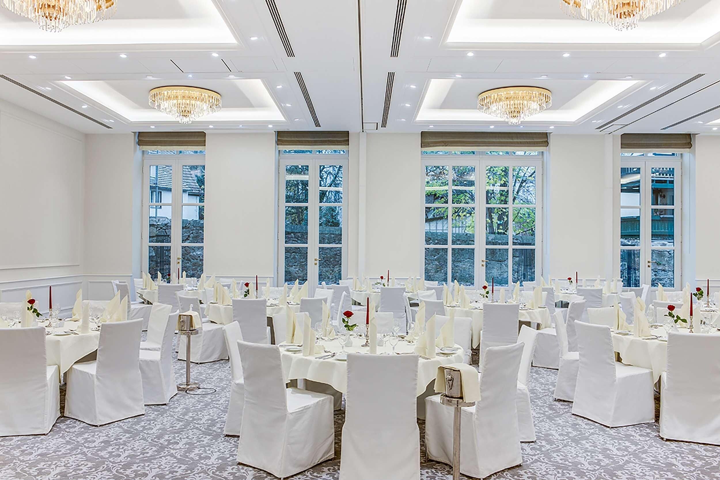Une salle de banquet avec des tables rondes, des chaises blanches, des lustres et de grandes fenêtres.
