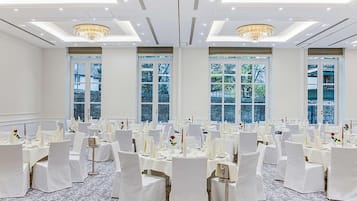 Une salle de banquet avec des tables rondes, des chaises blanches, des lustres et de grandes fenêtres.