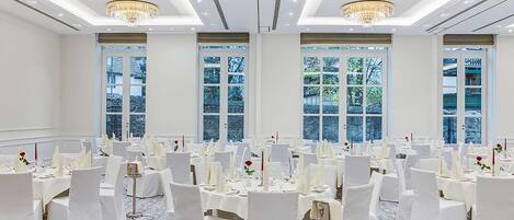 A banquet hall with round tables, white chairs, chandeliers, and large windows.