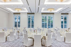 Un salón de banquetes con mesas redondas, sillas blancas, candelabros y ventanas amplias.