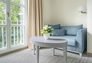 A room with a sofa, a round table, a potted plant, and a window with curtains. - Dorint Parkhotel Meissen (Nossen)