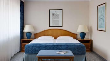 A hotel room with a double bed, two bedside tables with lamps, a framed document on the wall, and a small table with a magazine.