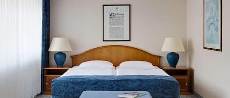 A hotel room with a double bed, two bedside tables with lamps, a framed document on the wall, and a small table with a magazine.