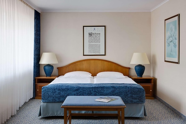Una habitación de hotel con cama doble, dos mesitas de noche con lámparas, un documento enmarcado en la pared y una pequeña mesa con una revista.
