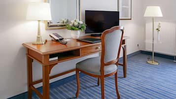 A hotel room with a wooden desk, a chair, a television, a telephone, a vase with flowers, and two lamps.