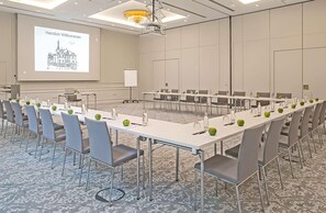 Ein Konferenzraum mit langen Tischen, Stühlen und einer Projektionsleinwand, auf der eine Begrüßungsnachricht angezeigt wird.