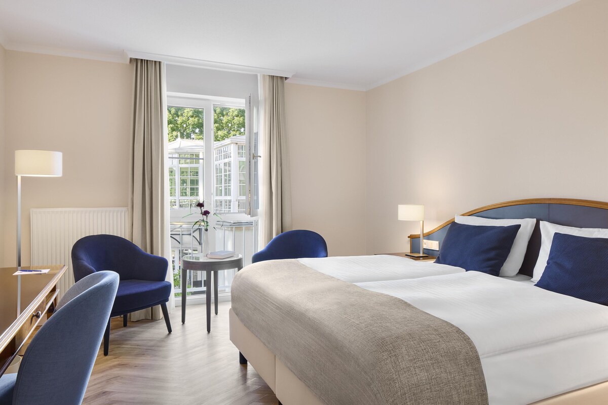 Une chambre d’hôtel avec un lit, deux chaises, une petite table et une fenêtre avec des rideaux.