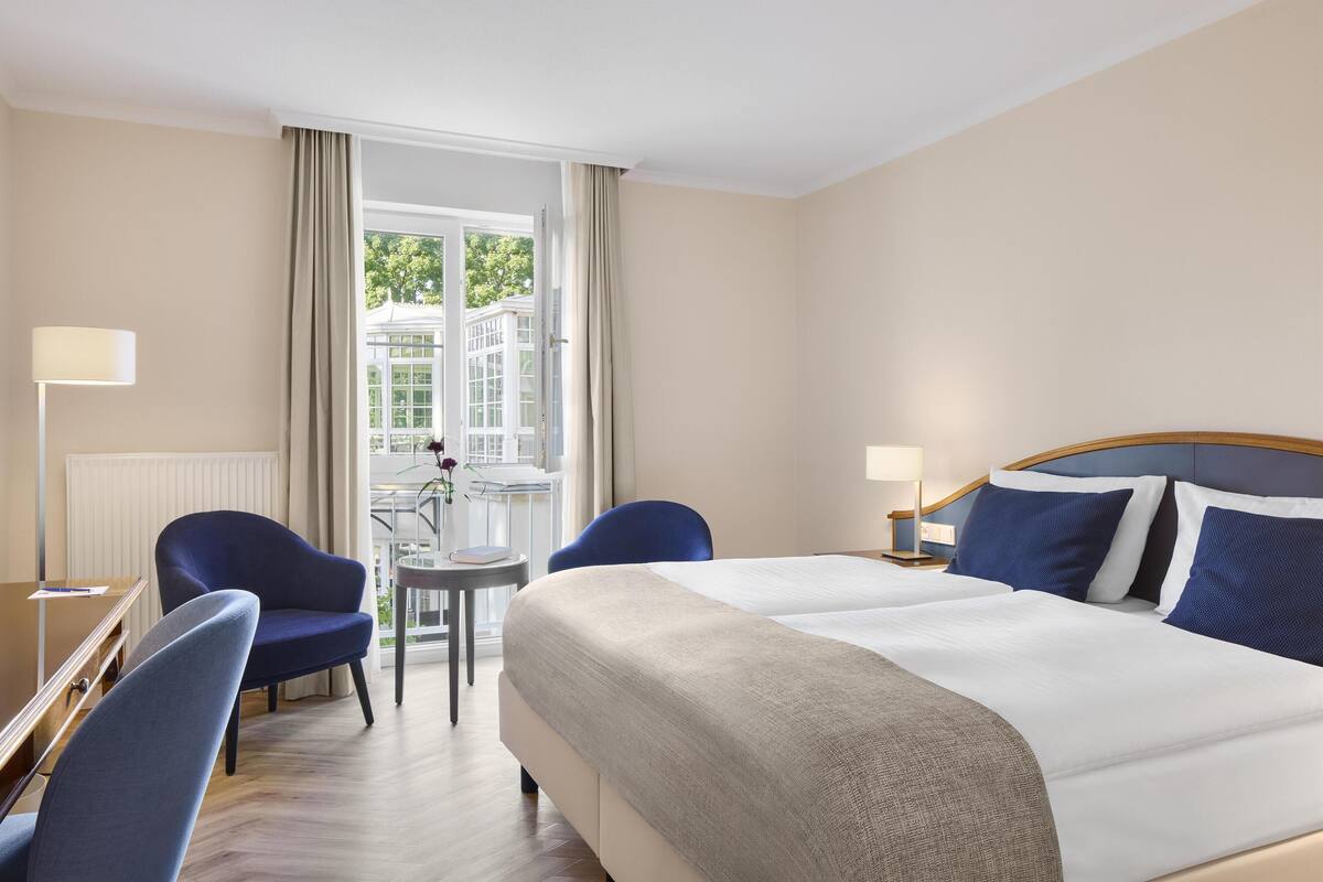 A hotel room with a bed, two chairs, a small table, and a window with curtains.