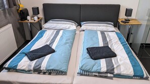 A double bed with striped bedding, two bedside tables with lamps, a radiator, and a small shelf with items. - Business-motel mit Xl-parking (Heimsheim)