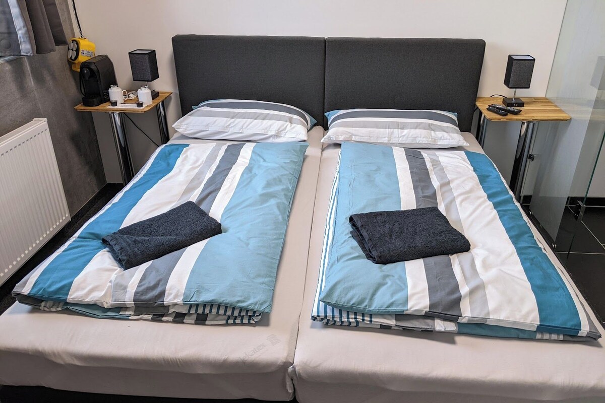 A double bed with striped bedding, two bedside tables with lamps, a radiator, and a small shelf with items.